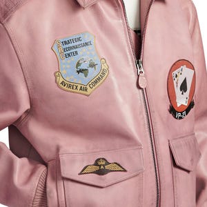 Pu&ograve; includere: Un giubbotto bomber in pelle rosa chiaro con chiusura frontale a zip e diverse toppe. Le toppe includono uno scudo blu con il testo Strategic Reconnaissance Center Avirex Air Command, una toppa circolare con carte da gioco e un emblema alato.