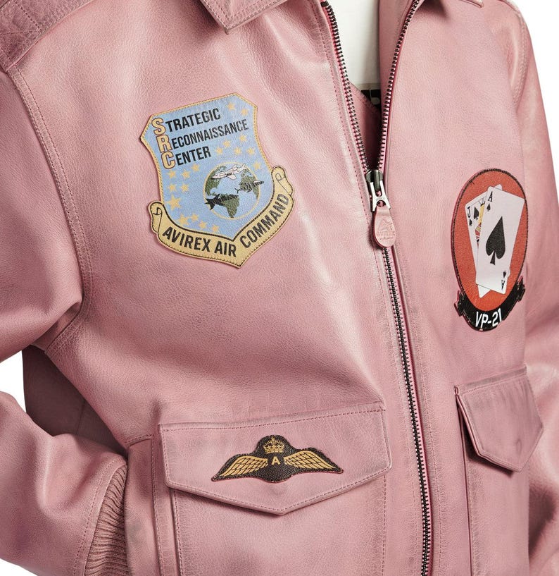 Pu&ograve; includere: Un giubbotto bomber in pelle rosa chiaro con chiusura frontale a zip e diverse toppe. Le toppe includono uno scudo blu con il testo Strategic Reconnaissance Center Avirex Air Command, una toppa circolare con carte da gioco e un emblema alato.