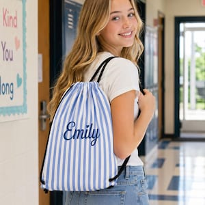 Peut inclure: Un sac à dos à cordon avec un motif à rayures verticales bleu clair et blanc. Le prénom Emily est imprimé en écriture cursive bleu foncé au centre du sac. Le sac possède des cordons noirs et est fabriqué dans un tissu léger.