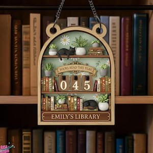 May include: Wooden bookshelf-shaped sign with a chain for hanging. The sign features a miniature library scene with books, plants, and a black cat. Text on the sign includes "BOOKS READ THIS YEAR", "045", and "EMILY'S LIBRARY".