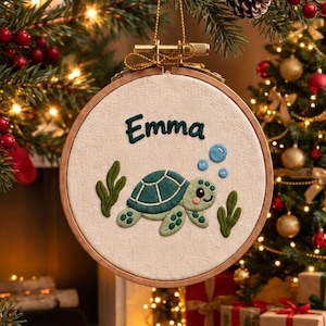 Pode incluir: Um bastidor de bordado circular de madeira contendo um painel de tecido branco com o nome Emma bordado em fio verde escuro acima de uma ilustração de tartaruga marinha, acompanhado por algas verdes e bolhas azuis, suspenso por um cordão dourado.
