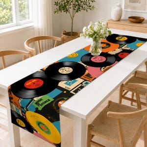 May include: A rectangular table runner featuring a vibrant, multi-colored graphic print of various vinyl records in black, yellow, orange, and pink, interspersed with abstract geometric shapes and text elements on a teal and black background.