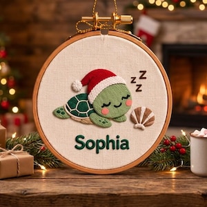 May include: A circular embroidery hoop containing a cream-coloured fabric with a stitched green turtle wearing a red and white Santa hat. The turtle is sleeping next to a brown seashell and the name Sophia is embroidered in dark green below the turtle.