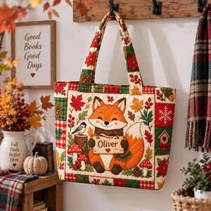 Può includere: Una borsa tote trapuntata con un'illustrazione centrale di una volpe sorridente che tiene un cartello in legno con la scritta Oliver e un piccolo cuore rosso. La borsa è decorata con motivi autunnali che includono funghi rossi e foglie verdi.