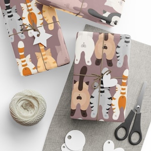 Puede incluir: Regalos envueltos con papel de regalo con temática de gatos. El papel presenta un patrón repetido de traseros de gatos en varios colores. Los regalos están atados con hilo y adornados con etiquetas en forma de estrella y corazón. También hay un ovillo de hilo, tijeras y etiquetas en blanco.