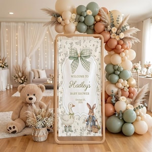 May include: A baby shower welcome sign with the text "Welcome to Hadley's Baby Shower October 25, 2026". The sign features a green plaid bow, a goose, and a rabbit. The sign is framed with a wooden border. A large teddy bear and balloon arch are also visible.