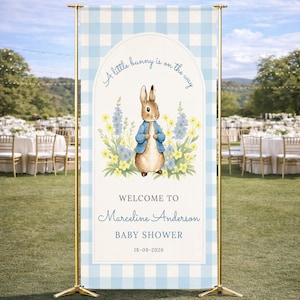Może przedstawiać: Witamy znak na baby shower z ilustracją królika w niebieskiej kurtce, otoczonego kwiatami. Znak ma niebiesko-biały wzór w kratkę i głosi "A little bunny is on the way" i "Welcome to Marceline Anderson Baby Shower 18-09-2026."