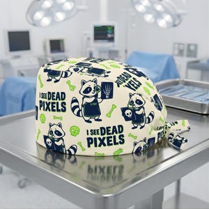 May include: A cream-colored surgical cap featuring a repeating pattern of raccoons, bones, and skulls. The raccoons hold X-ray images and skulls. Text on the cap reads "I SEE DEAD PIXELS" and "DEAD YELS".