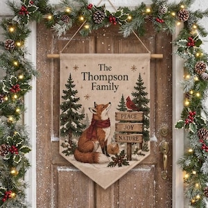 May include: A beige fabric wall hanging banner featuring a fox wearing a red scarf, pine trees, and a wooden sign with the text The Thompson Family, Peace, Joy, Nature. The banner is suspended from a wooden rod with a string.