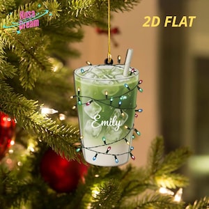 Könnte beinhalten: Ein 2D-flacher Weihnachtsschmuck in Form eines grünen Getränks mit Eis und Strohhalm, umwickelt mit bunten Lichterketten. Der Name "Emily" steht auf dem Glas. Der Schmuck hängt an einem goldenen Faden vor einem Weihnachtsbaumhintergrund.