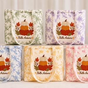 May include: Five quilted tote bags in green, purple, blue, yellow, and pink, each featuring a white background with a floral pattern. Centered on each bag is an embroidered stack of three pumpkins with autumn leaves and the text Hello Autumn.