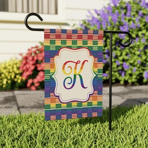 May include: A garden flag with a colourful design featuring the letter "K" in rainbow colours. The flag has a cream-coloured frame around the letter and is set against a background of rainbow stripes and American flag patterns. The flag is displayed on a black metal stand.