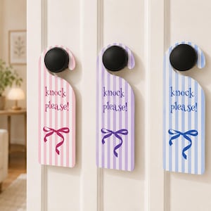 Puede incluir: Tres colgadores de puerta verticales en rosa, lavanda y azul claro, cada uno con rayas verticales blancas, el texto "knock please!" en un color a juego y un gráfico de lazo en la parte inferior, colgados de pomos de puerta negros.
