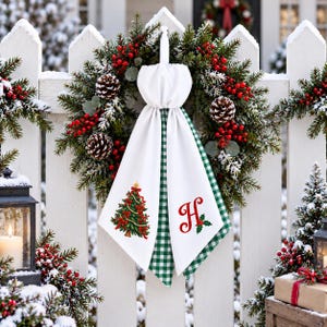 May include: A Christmas wreath featuring green pine branches, red berries, and pinecones, adorned with a white fabric hanging towel. The towel displays a printed Christmas tree on one side and a red letter H with holly on the other.