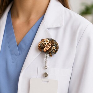 Könnte beinhalten: Ein Ausweishalter mit einem gehirnförmigen Ornament, das mit metallischen Zahnrädern, Getriebeteilen und kleinen weißen Blumen verziert ist. Das Gehirn ist in Braun- und Beigetönen gehalten und an einer transparenten Kunststoffrolle befestigt.