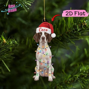 Puede incluir: Un adorno colgante plano y bidimensional que muestra a un perro marrón y blanco con un gorro de Papá Noel rojo con el nombre Coco impreso en letras blancas. El perro está envuelto en una cadena de luces navideñas de colores.