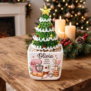 May include: A white square ceramic planter featuring the name Olivia and a colourful baking-themed graphic with a pink stand mixer, rolling pin, and cookies. A green artificial Christmas tree with a yellow star topper sits inside the planter.
