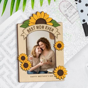 Może przedstawiać: Drewniana ramka na zdjęcia z akcentami słoneczników i napisem "BEST MOM EVER" i "HAPPY MOTHER'S DAY". Ramka zawiera zdjęcie matki i córki. Ramka jest ozdobiona żółtymi i brązowymi słonecznikami.