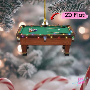 May include: A 2D flat hanging ornament shaped like a wooden pool table with a green felt surface, billiard balls, and two cues. The table is decorated with a string of colorful Christmas lights and hangs from a gold cord against a blurred background.