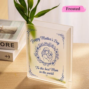Puede incluir: Un jarrón de acrílico esmerilado con forma de libro, con un diseño floral azul y el texto "Happy Mother's Day". El jarrón presenta una ilustración de una madre y un niño y el mensaje "To the best Mom in the world."