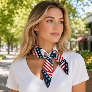 May include: A square neck scarf featuring a patriotic pattern of red and white stripes combined with a dark blue border adorned with white stars. The scarf is tied in a simple knot at the neck, displaying a geometric arrangement of the flag-inspired design.