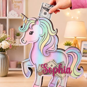 Pu&ograve; includere: Un salvadanaio in legno a forma di unicorno dai colori pastello, con corno, criniera e coda nelle tonalit&agrave; del rosa, blu, giallo e verde. Il salvadanaio ha una fessura sulla parte superiore per inserire il denaro e il nome "Sophia" &egrave; visualizzato sulla parte anteriore. Una mano inserisce una banconota da 100 &euro;.