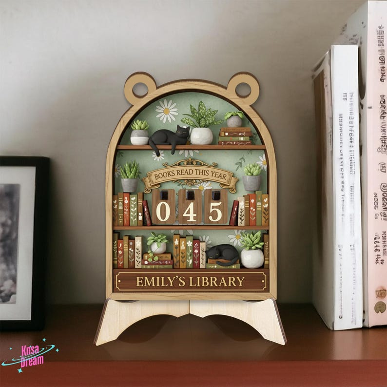 May include: Wooden bookshelf-shaped decor with a bear-ear top. It features miniature books, plants, and a black cat figurine. The text "BOOKS READ THIS YEAR" and "045" are displayed. The bottom reads "EMILY'S LIBRARY".