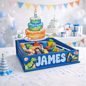 May include: A blue wooden toy box with the name "JAMES" in white letters. The box is filled with wooden blocks, toy cars, and stuffed animals. A colorful birthday cake cutout and a party hat are also visible.