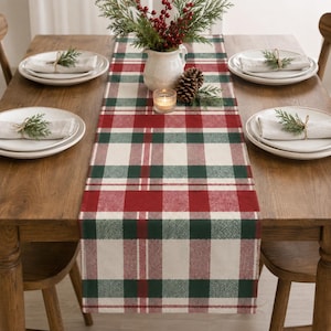 May include: A rectangular table runner featuring a red, dark green, and cream plaid pattern. The runner is centered on a wooden table with four white plates, each topped with a folded napkin and a small sprig of greenery, plus a central vase.