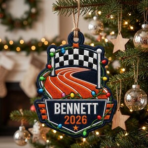 Op de afbeelding: Een donkerblauw, schildvormig kerstornament met een geblokte finishvlag, een oranje hardloopbaan en een snoer met gekleurde kerstverlichting. De tekst BENNETT en het jaartal 2026 staan in witte en oranje letters op het ornament.