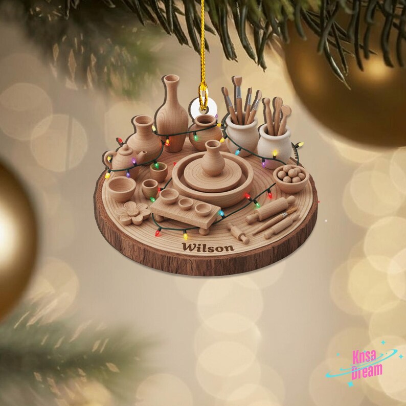 May include: A wooden Christmas ornament featuring a pottery theme. The ornament includes miniature pottery tools, a pottery wheel, and various pottery pieces. The name "Wilson" is inscribed on the ornament, which is decorated with colorful lights.
