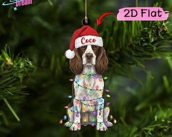 May include: A flat, two-dimensional hanging ornament featuring a brown and white spaniel dog wearing a red Santa hat with the name Coco printed on the brim. The dog is wrapped in a string of colorful, glowing Christmas lights.