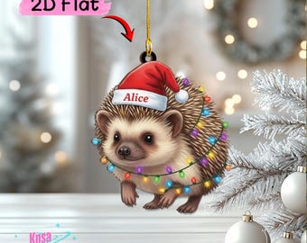 May include: A 2D flat hanging ornament featuring a brown hedgehog wearing a red Santa hat with the name Alice printed on the white brim. The hedgehog is wrapped in a string of colorful Christmas lights and hangs from a gold cord.