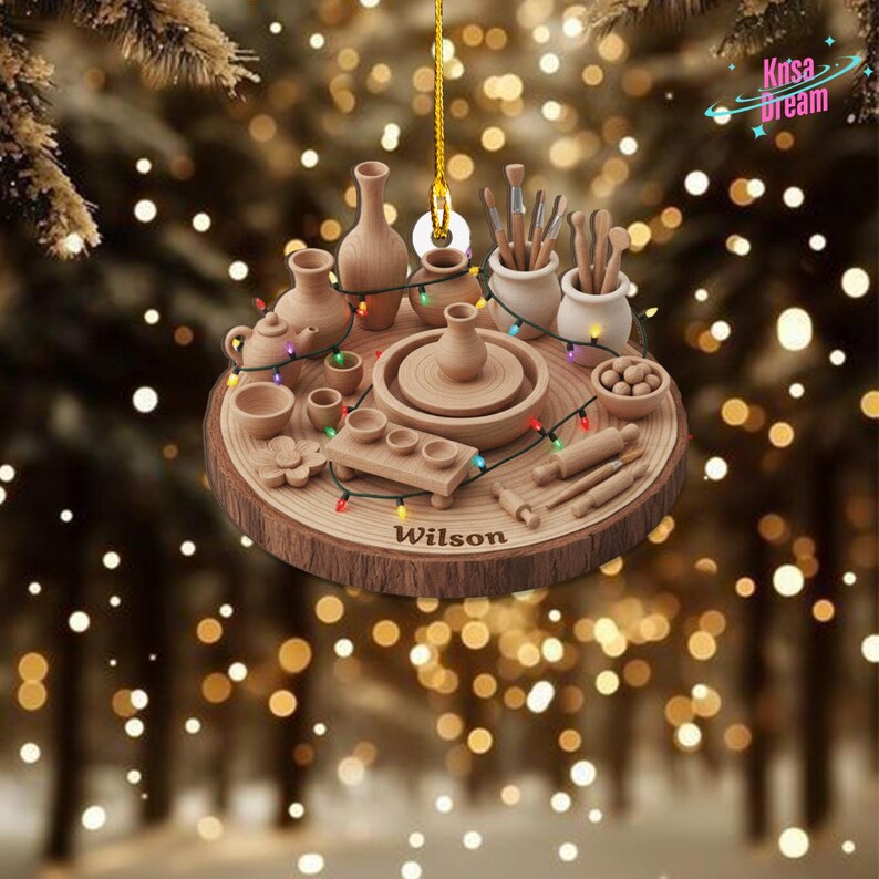 May include: Wooden ornament featuring pottery tools and miniature pottery items, including a pottery wheel, vases, and brushes. The ornament is shaped like a wood slice and includes the name "Wilson". Decorative lights are strung around the pottery items.