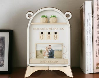May include: A white wooden wedding countdown calendar with the text "DAYS UNTIL WE SAY I DO" and the numbers "089". It features a photo frame with a couple's picture and the names "EMILY & DAVID". Two small potted succulents sit on a shelf above.