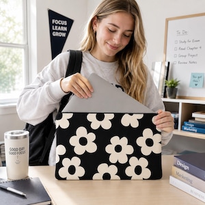 May include: A rectangular laptop sleeve featuring a repeating pattern of large, cream-colored flowers with dark centers against a solid black background. The sleeve is constructed from a soft, flexible material and is designed to hold a thin, silver laptop.