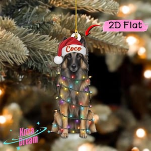 May include: A 2D flat hanging ornament featuring a German Shepherd dog wearing a red and white Santa hat with the name Coco printed on the brim. The dog is wrapped in a string of colorful Christmas lights. A pink label reads 2D Flat.