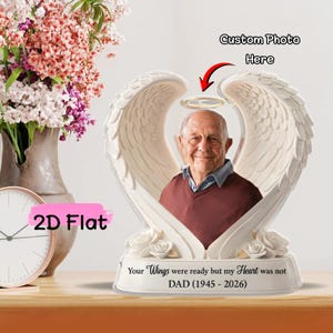 Könnte beinhalten: Eine Gedenkfigur mit Engelsflügeln in Weiß und einem Foto einer Person. Die Flügel umrahmen das Foto. Auf dem Sockel steht der Text "Your Wings were ready but my Heart was not" und die Daten 1945-2026. Der Text "Custom Photo Here" ist ebenfalls sichtbar.