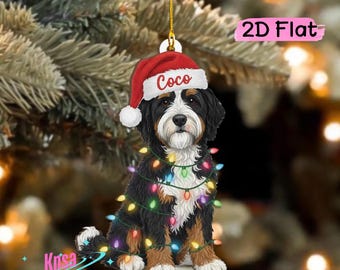 Op de afbeelding: Een platte 2D-hangdecoratie met een zwart, wit en bruin hondje dat een rood-witte kerstmuts draagt met de naam Coco op de rand. De hond is omwikkeld met een snoer van kleurrijke, gloeiende kerstverlichting en heeft een gouden ophangkoord.