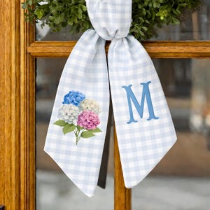 Op de afbeelding: Lichtblauw en wit geruit lint met een bloemenhortensia-ontwerp en de letter "M". Het lint is om een groene krans gebonden en hangt aan een houten deur.