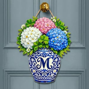 May include: A wooden door hanger featuring a blue and white vase with the letter "M" and a floral arrangement of pink, white, and blue hydrangeas, with green leaves. The hanger is suspended by a rope and a gold-colored knob.