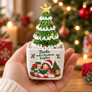 May include: A small square white ceramic planter featuring a green Christmas tree with a gold star topper. The planter displays the text "Books make Christmas brighter" above an illustration of a person reading books with a cat and a lantern.