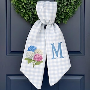 Puede incluir: Bufanda para puerta a cuadros azules y blancos con un diseño floral de hortensias y la letra "M". La bufanda cuelga de una puerta azul, con una corona verde en el fondo.