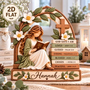 May include: A decorative figurine featuring a person with long brown hair reading a Holy Bible, framed by an arch with white flowers and green leaves. A wooden cross sits above a stack of books labelled with religious text and the name Hannah.