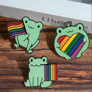 Puede incluir: Tres pines de esmalte con ranas de color verde claro. Una rana sostiene una bandera arcoíris horizontal, otra un corazón con los colores del arcoíris y la tercera rana sostiene una bandera arcoíris vertical. Tienen ojos y boca de color negro.