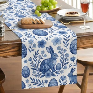 May include: A white table runner with a blue floral and Easter-themed design. The runner features blue Easter eggs, bunnies, and flowers. The runner is on a wooden table with food and drinks.