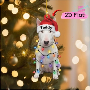 May include: A 2D flat hanging ornament featuring a white Bull Terrier dog wearing a red Santa hat with the name Teddy printed on the brim. The dog is wrapped in a string of colorful Christmas lights with red, blue, yellow, and purple bulbs.
