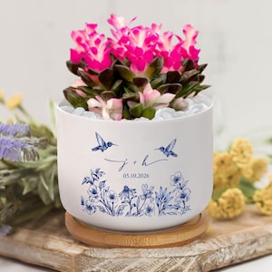 Puede incluir: Una maceta de cerámica blanca con una vibrante planta de flores rosas. La maceta presenta diseños de colibríes y flores azules, junto con las iniciales "j + h" y la fecha "05.10.2026". La maceta se encuentra sobre una base redonda de madera.