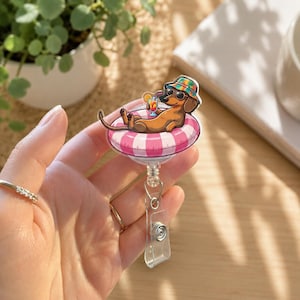 Pode incluir: Um porta-crachá com um cão basset castanho a usar um chapéu colorido e óculos de sol, deitado numa boia insuflável às riscas cor-de-rosa e brancas enquanto segura uma bebida tropical com uma palhinha. Fixado a um clipe de plástico transparente.