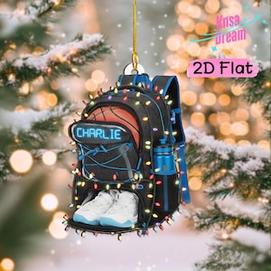 Puede incluir: Un adorno colgante plano 2D con forma de mochila negra con un gráfico de baloncesto, el nombre CHARLIE en texto azul, una botella de agua azul y zapatillas blancas, todo envuelto en luces navideñas de colores con un cordón dorado.
